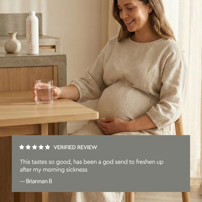A pregnant woman in a beige linen dress sits at a wooden table, holding a glass of water. A white mouthwash bottle labeled MamaSmiles is visible on a shelf in the background.