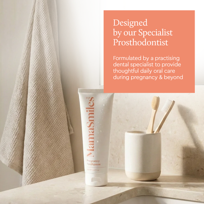 A white tube of MamaSmiles Pregnancy Toothpaste sits on a bathroom counter beside a toothbrush holder with two wooden toothbrushes and a hanging towel. 