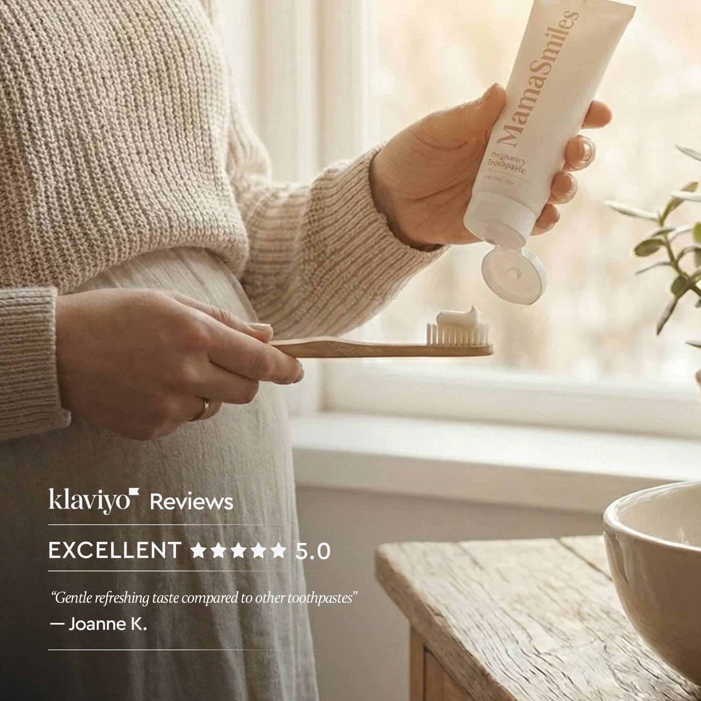 A person in a beige knit sweater holds a tube of MamaSmiles toothpaste while squeezing white toothpaste onto a wooden toothbrush, with a soft window background and a review quote overlaid on the left.