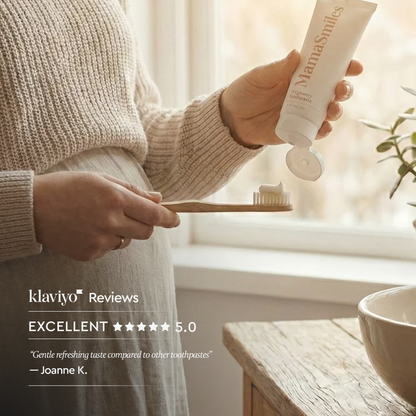 A person in a beige knit sweater holds a tube of MamaSmiles toothpaste while squeezing white toothpaste onto a wooden toothbrush, with a soft window background and a review quote overlaid on the left.